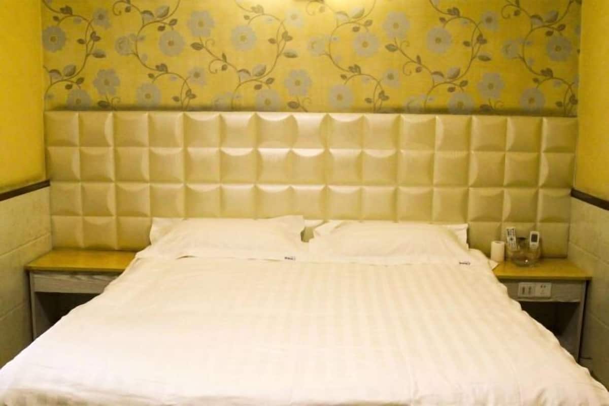 Une chambre d’hôtel avec une tête de lit capitonnée, un lit avec du linge de maison blanc, une table de chevet avec une lampe et un papier peint à motifs floraux.