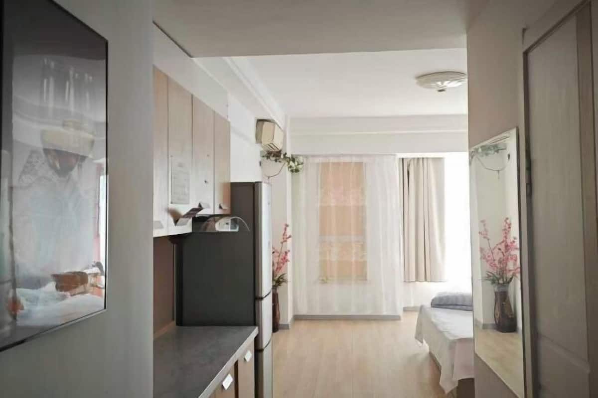 A hotel room with a bed, a small kitchenette, a bathroom, and a large window with sheer curtains.