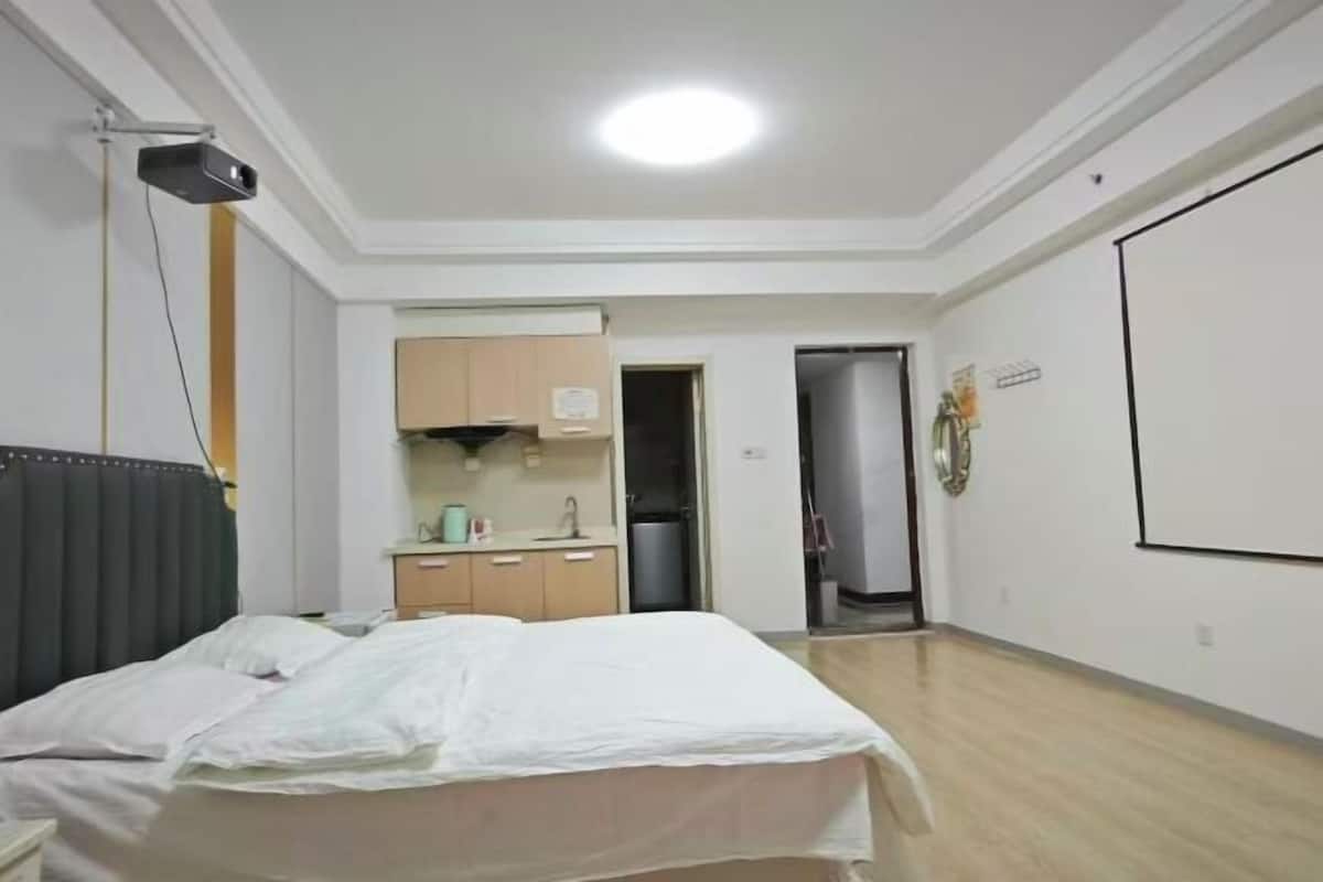 A bedroom with a bed, a desk, a kitchenette, and a door leading to another room.