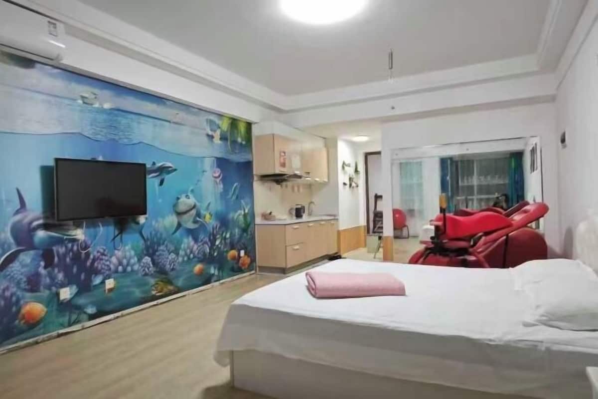 A hotel room with a bed, a television, a mural of an underwater scene, a kitchenette, and a red chair.
