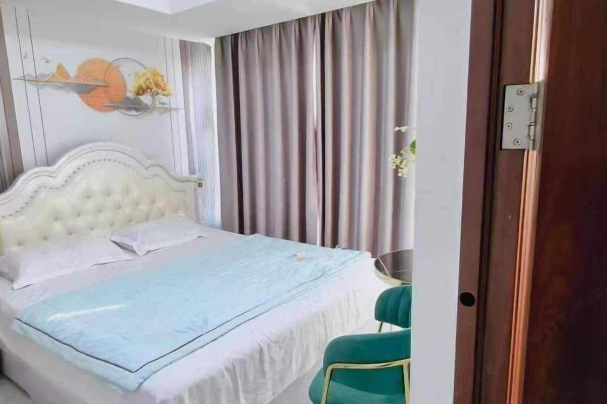 A bedroom with a tufted headboard, a bed with white linens, a teal chair, and a view of a curtain.