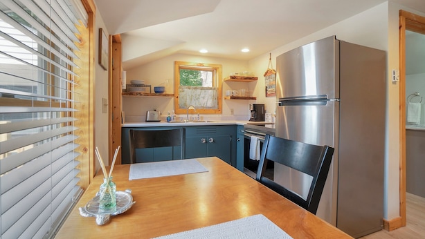 Une cuisine avec une table en bois, des chaises, un réfrigérateur en acier inoxydable et une fenêtre avec des stores.