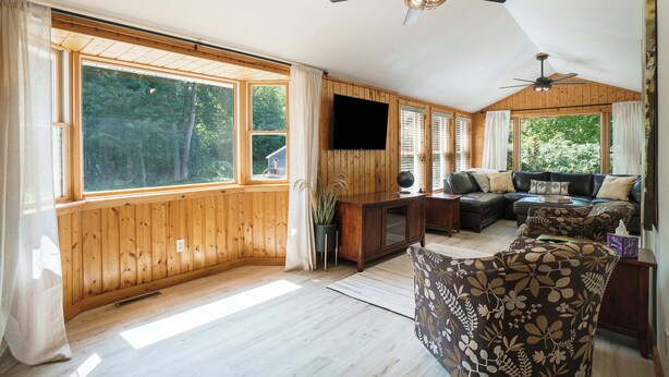 Ein Zimmer mit Holzwänden, einem Deckenventilator, großen Fenstern, einer Couch und einem Fernseher.