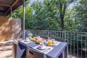 An outdoor dining area with a table set for breakfast, surrounded by a wooden deck and metal railing, overlooking a forested area.