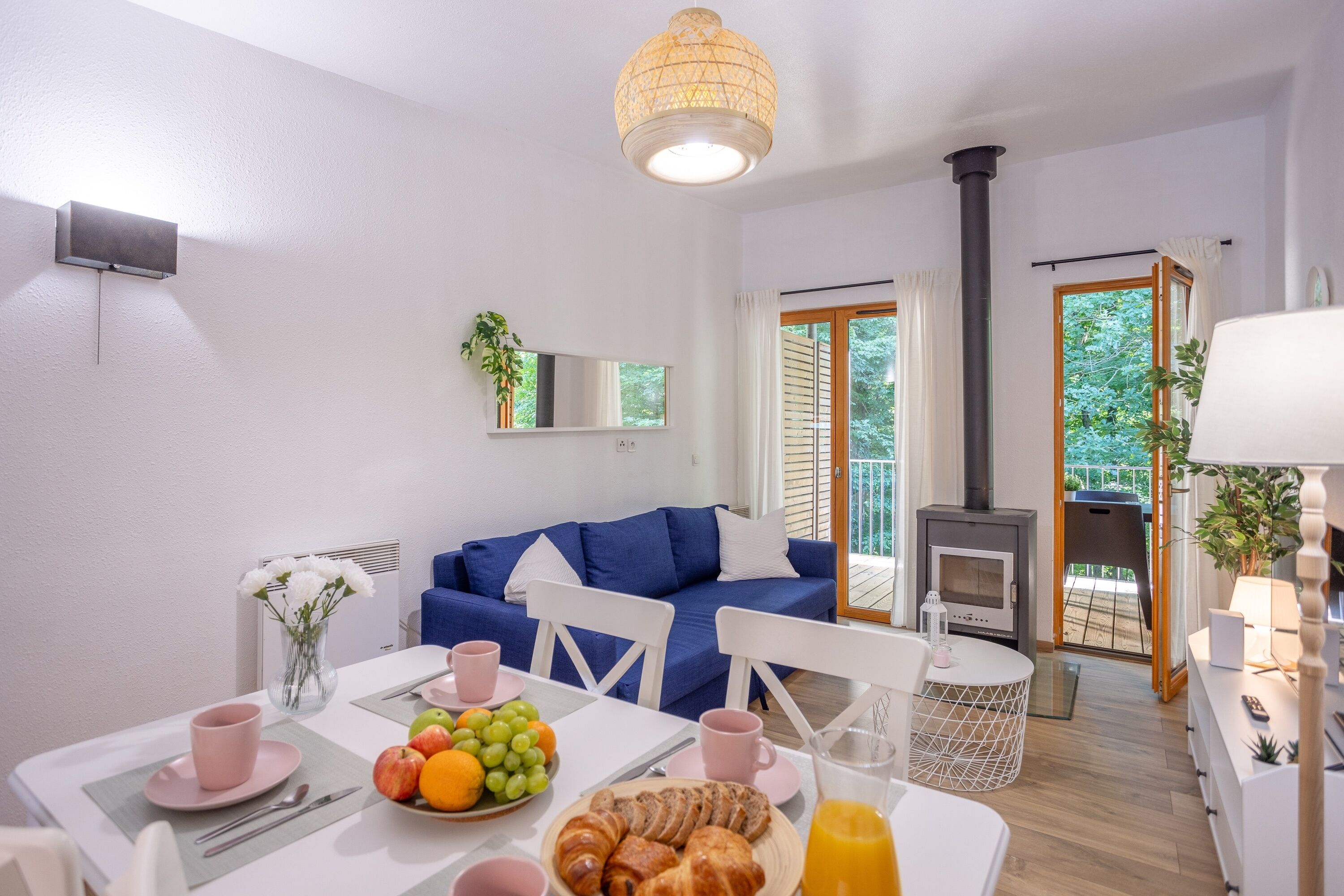 Un salon moderne avec un canapé bleu, une table à manger où sont disposés des aliments pour le petit-déjeuner, une cheminée et un balcon offrant une vue agréable.