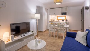 Un espacio habitable compacto con televisor de pantalla plana, una mesa de centro blanca, un sofá azul, una zona de comedor con una mesa y sillas blancas, y una kitchenette con microondas y un refrigerador pequeño.