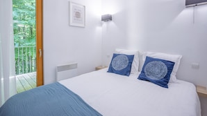 Un dormitorio con cama, dos almohadas, una lámpara de pared, una cita en un marco y una ventana con vista a follaje.