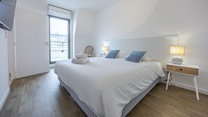 A bedroom with a large bed, a bedside table with a lamp, a chair, and a view of the outside through a window. - Le Vertigo - Appt proche de la plage (Dinard)