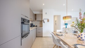 A modern kitchen with built-in appliances, a dining area with a wooden table and chairs, and a hanging pendant light. - Le Vertigo - Appt proche de la plage (Dinard)