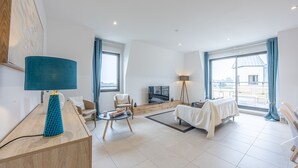 A modern living room with a sofa, a coffee table, a TV, and a large window with curtains. - Le Vertigo - Appt proche de la plage (Dinard)