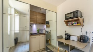 A compact kitchen with a microwave, toaster, and kettle on a shelf, a sink, and a small dining area with a chair. - Cozy Corner - Appt avec jardin pour 4 (Tournan-en-Brie)