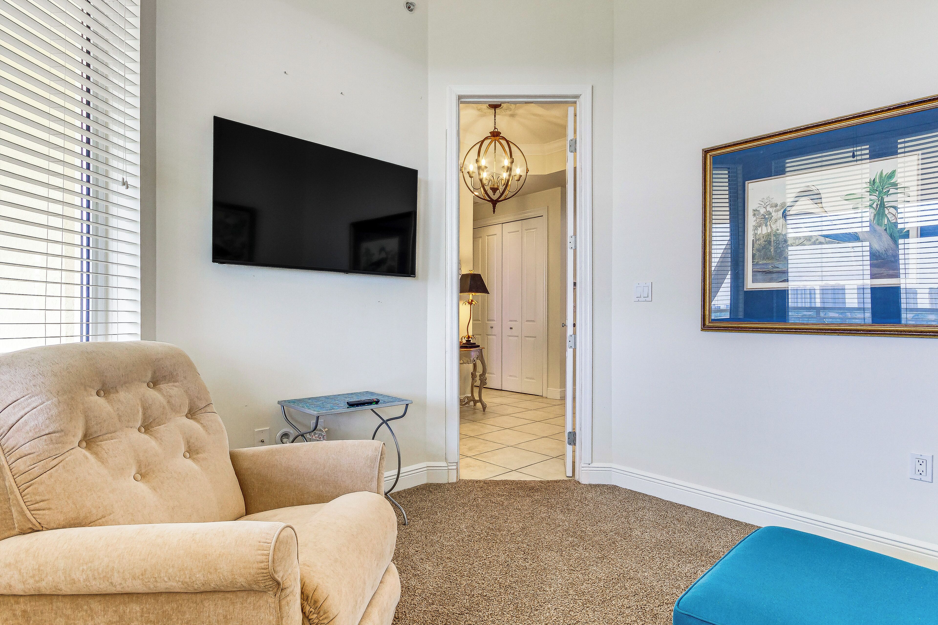 A living room with a beige sofa, a TV mounted on the wall, a chandelier, and a framed picture.