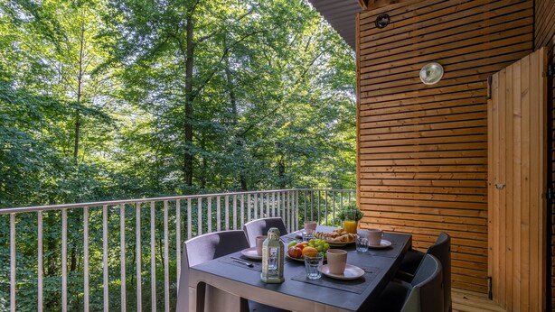 Eine Holzterrasse mit einem für das Frühstück gedeckten Tisch, umgeben von Stühlen, mit Blick auf ein bewaldetes Gebiet.