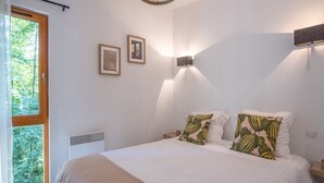 Ein Schlafzimmer mit großem Fenster, einem Bett mit weißer Bettwäsche, zwei Wandleuchten und zwei gerahmten Bildern an der Wand.