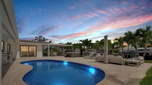 A swimming pool with a curved edge, surrounded by lounge chairs and palm trees, under a sunset sky.