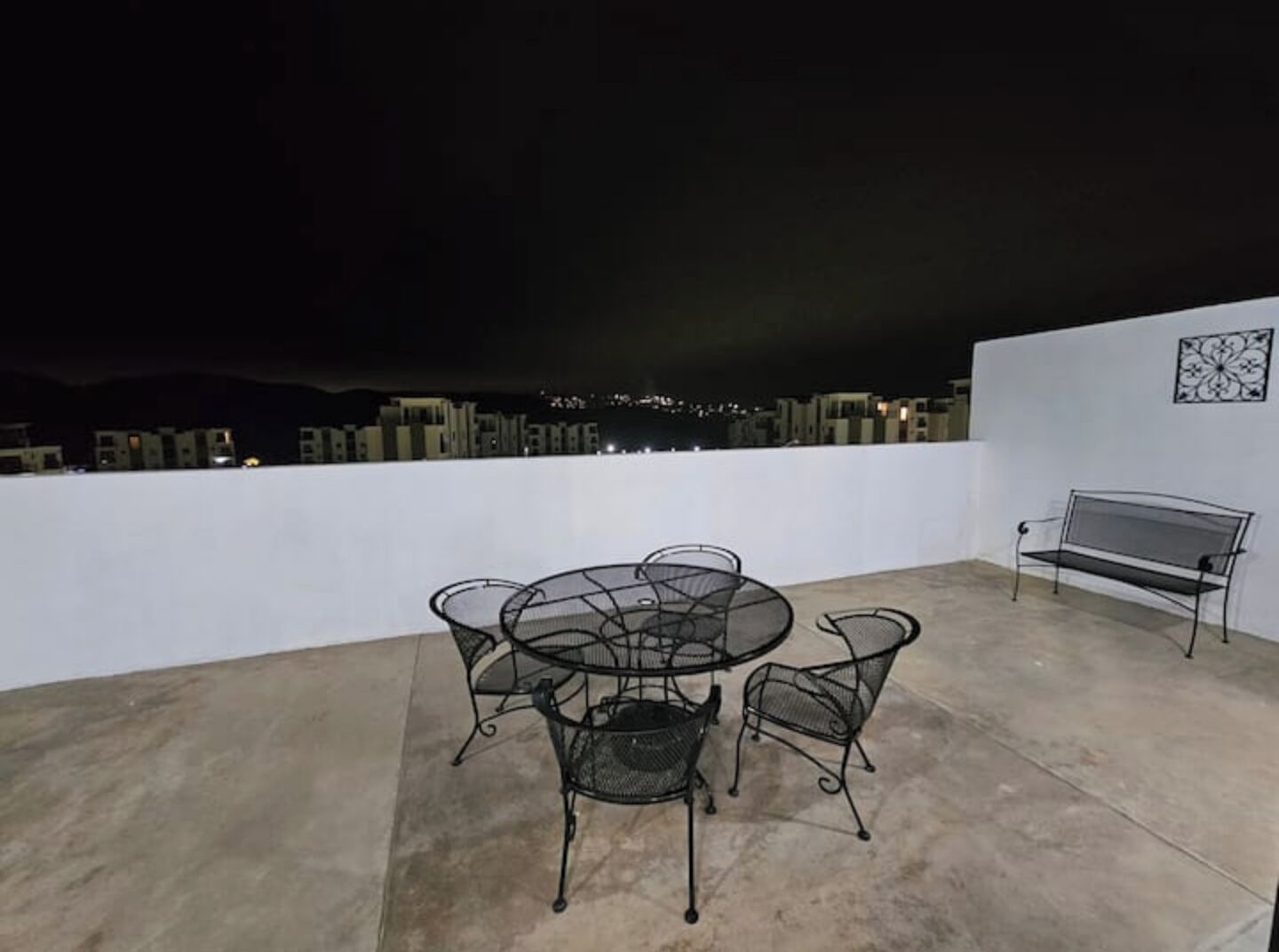 A rooftop terrace with a round table and chairs, a bench, and a city view at night.