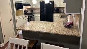 Una cocina con armarios blancos, encimera de granito, refrigerador negro, estufa y microondas. Hay dos lámparas colgantes sobre la encimera.
