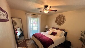 Un dormitorio con cama, ventilador de techo, ventana con cortinas, mesita de noche con lámpara y un adorno en la pared.