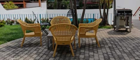 Un espace extérieur aménagé avec une surface pavée, trois chaises en osier, un barbecue et une clôture blanche.