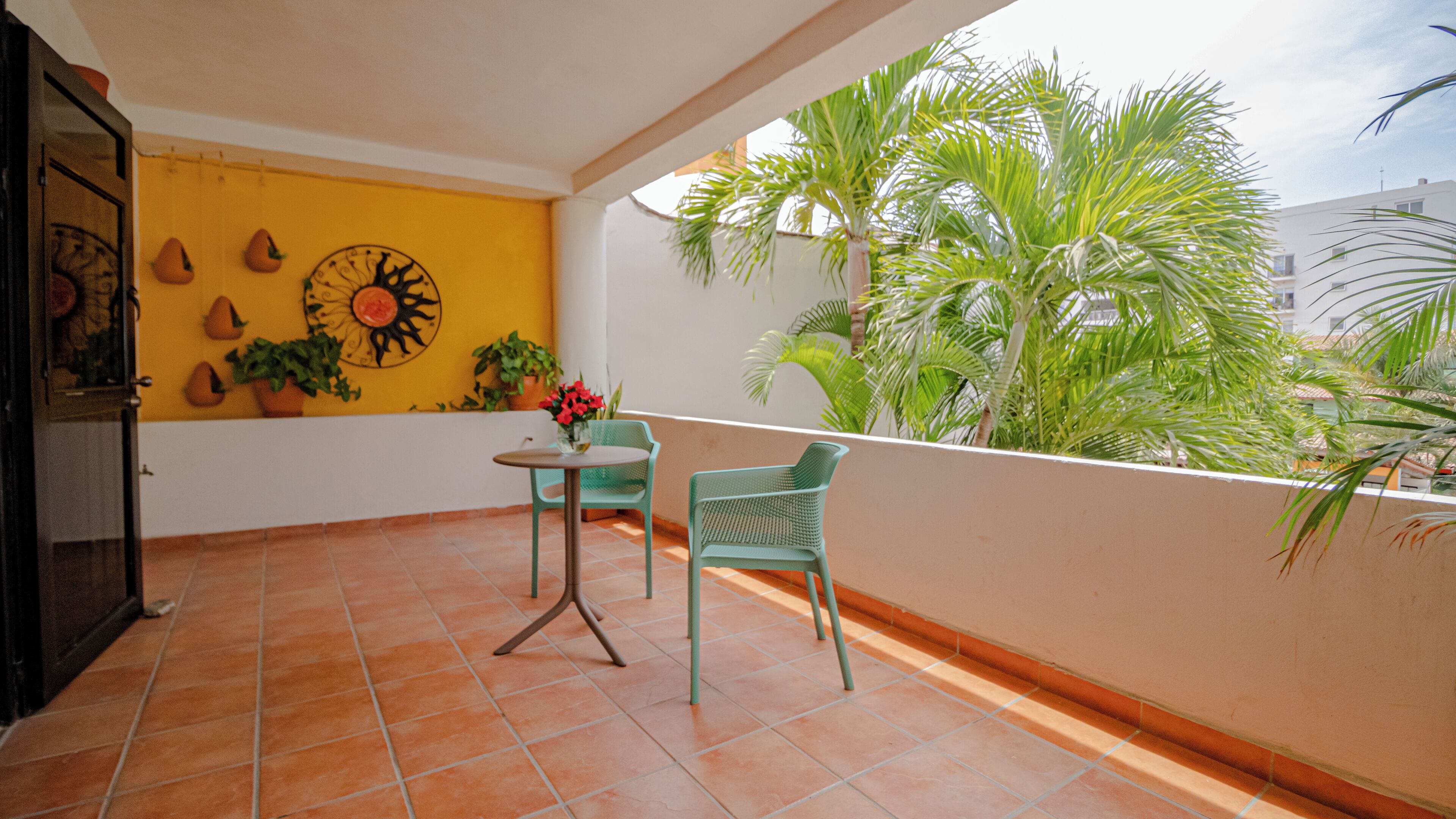 A balcony with terracotta tiles, a small round table, two chairs, potted plants, and a wall with a decorative sun motif.