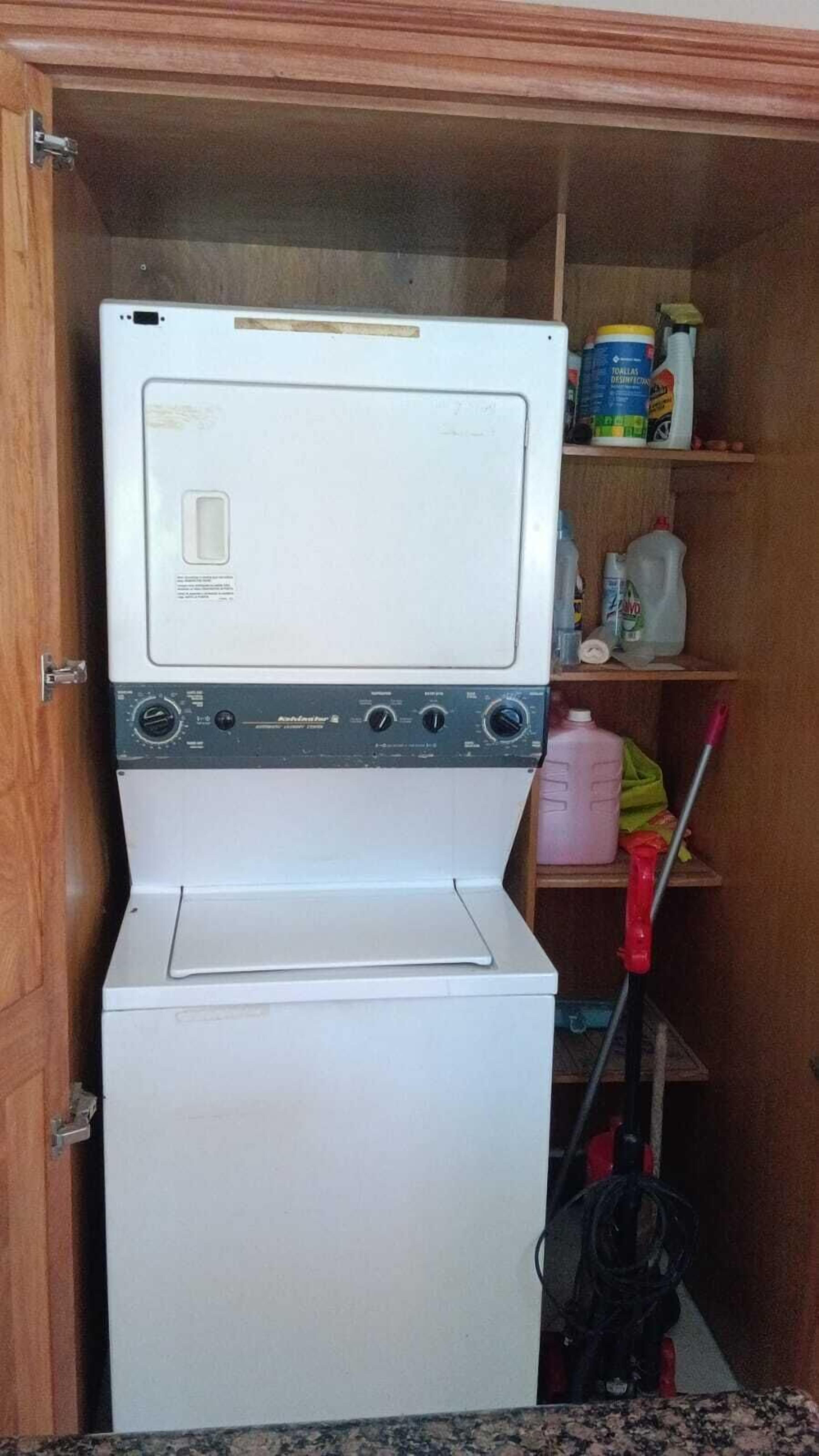 Een kleine wasruimte met een gestapelde wasmachine en droger, een plank met schoonmaakmiddelen, en een dweil op de vloer.