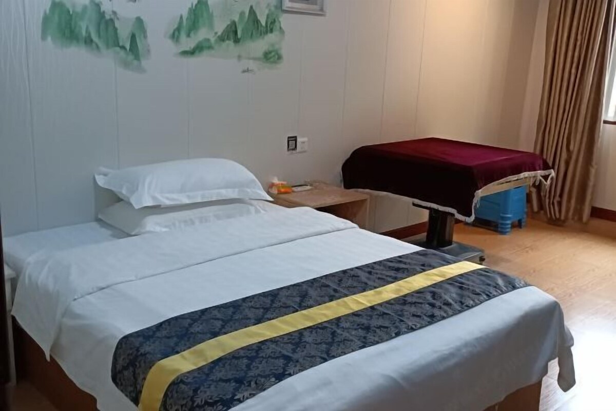 A neatly made bed with a white and blue bedspread, a wooden nightstand with a red cloth, and a wall with a green landscape painting.