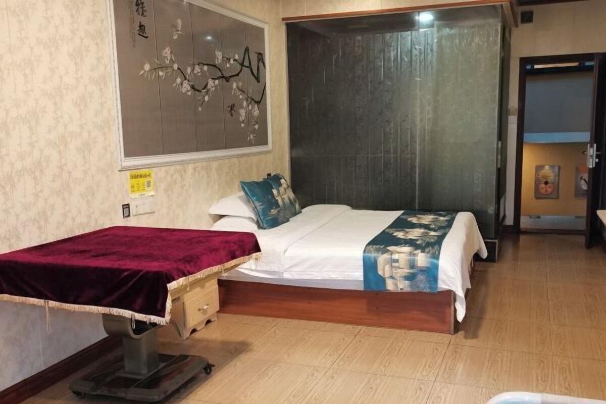 A bedroom with a bed, a massage table, a wooden floor, and a wall with a floral painting.