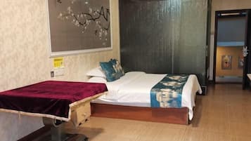 A bedroom with a bed, a massage table, a wooden floor, and a wall with a floral painting.