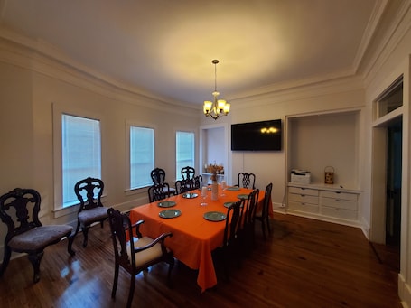 A dining room with a wooden table, chairs, a chandelier, and a TV mounted on the wall.