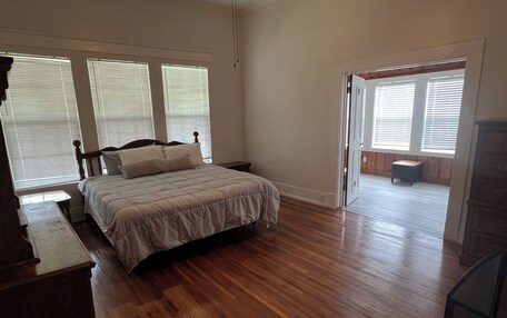 A bedroom with a wooden floor, a bed with a headboard, and a nightstand. There is a door leading to a balcony with a view of the outside.