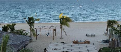 Uma praia com cabanas de palha, palmeiras e uma área arenosa com guarda-sóis e assentos.