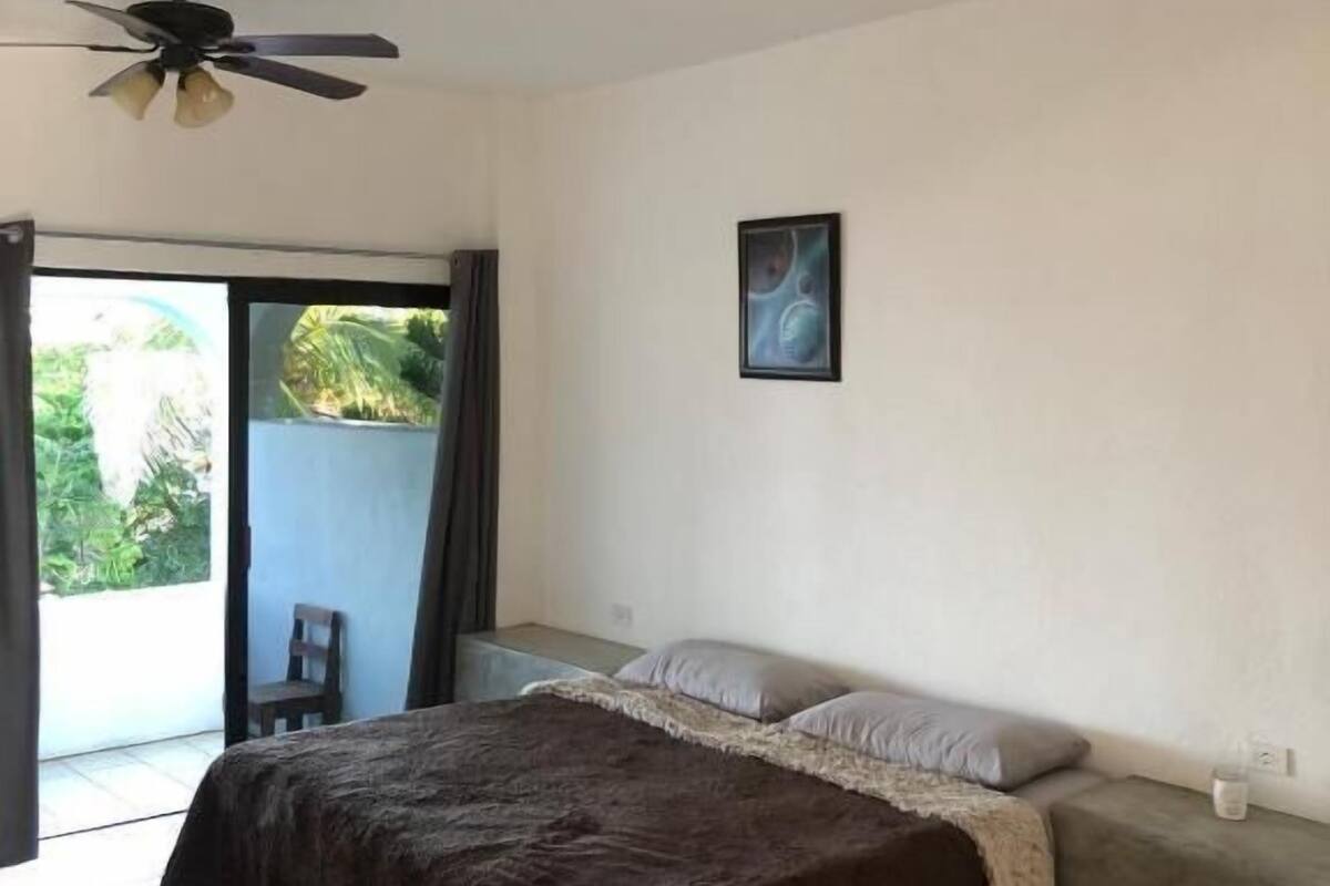 Une chambre à coucher avec un lit, un ventilateur de plafond, un tableau au mur et une vue sur l’extérieur grâce à une porte coulissante.