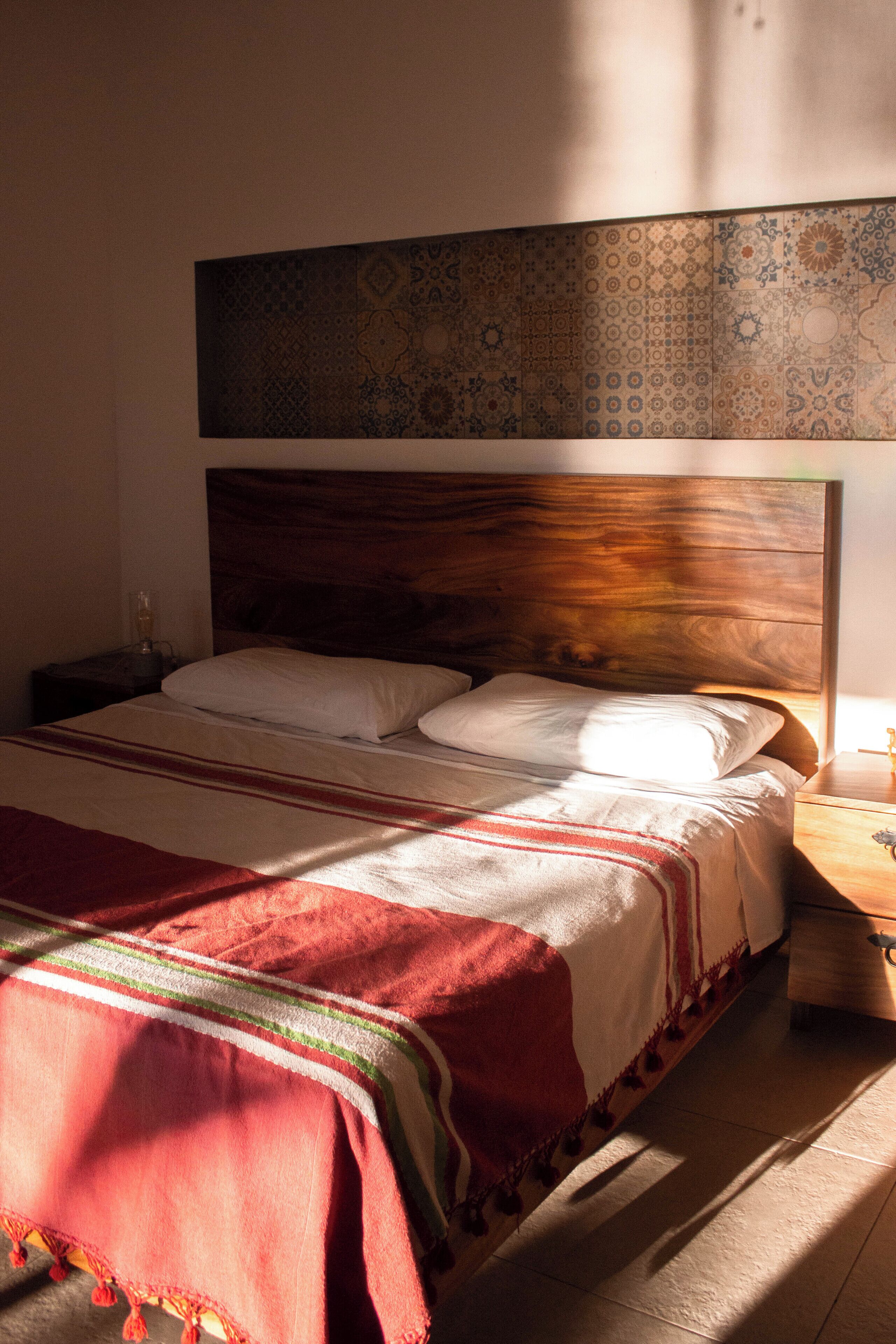 Una cama bien tendida con un cabecero estampado, una mesita de noche con una lámpara y una colcha roja y blanca.