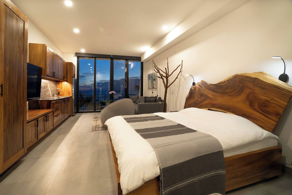 Une chambre moderne avec un grand lit, une tête de lit en bois et une vue sur le paysage urbain, visible à travers la porte-fenêtre coulissante.