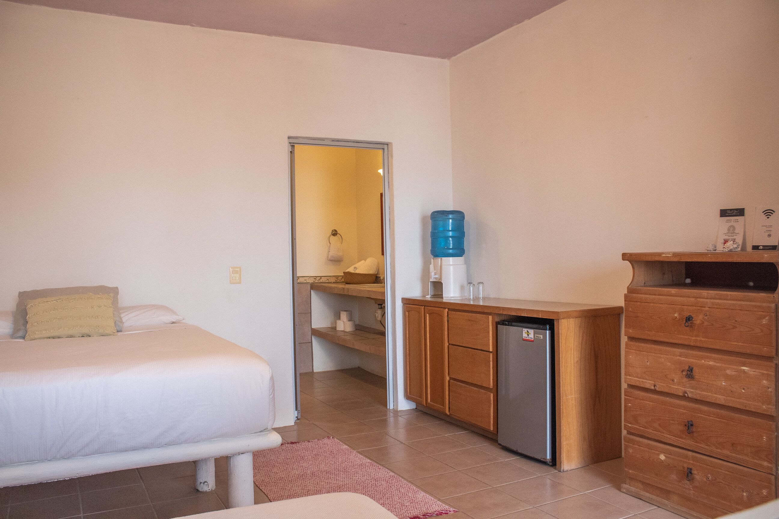 A compact hotel room with a bed, a small kitchenette, a water dispenser, and a wooden dresser.