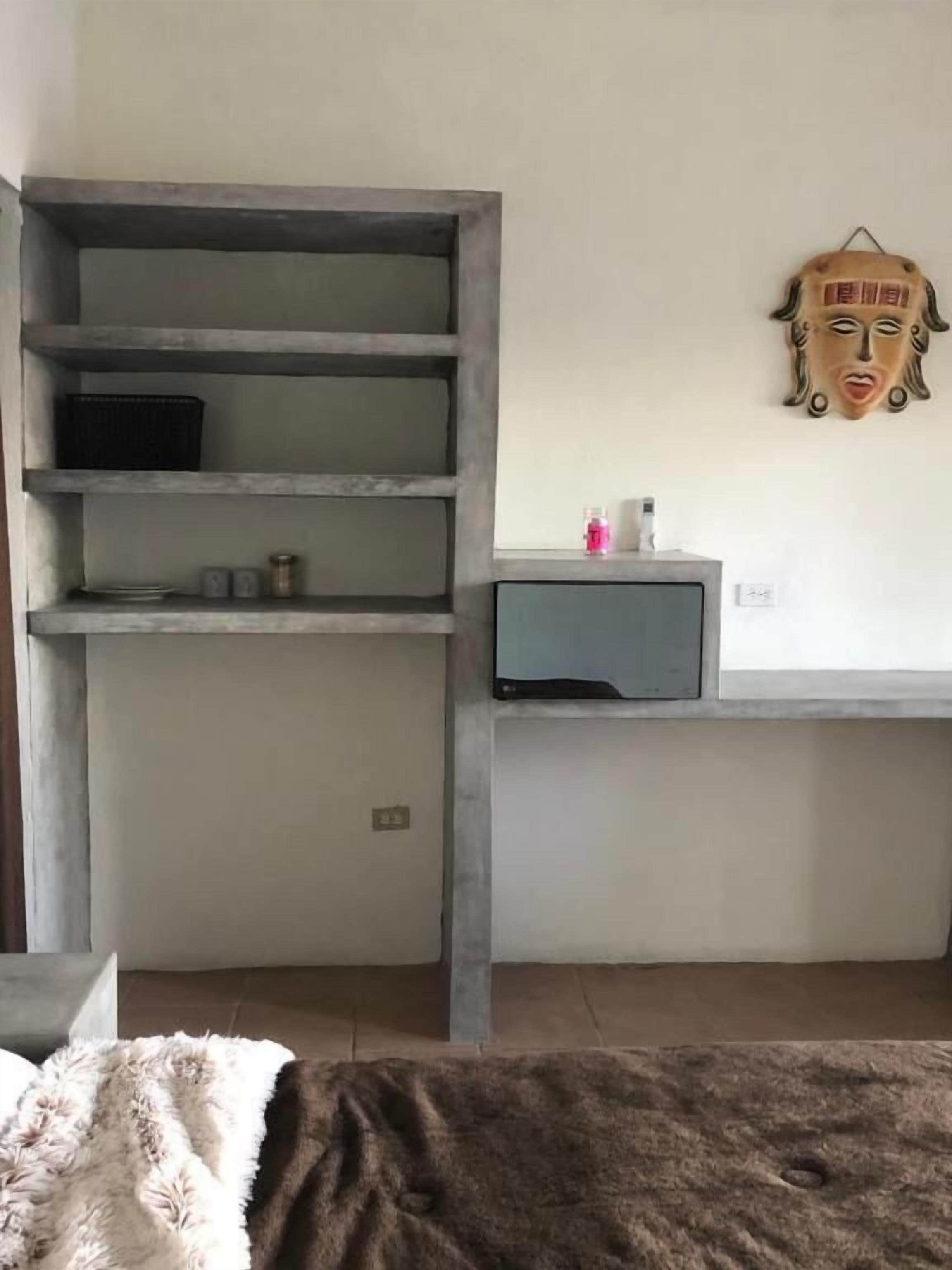 A bedroom with a bed, a grey wardrobe, a small table with a bottle, and a decorative mask on the wall.