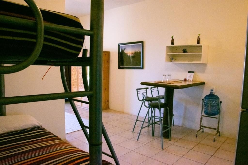 A dormitory room with bunk beds, a desk, chairs, a water dispenser, and a framed picture on the wall.