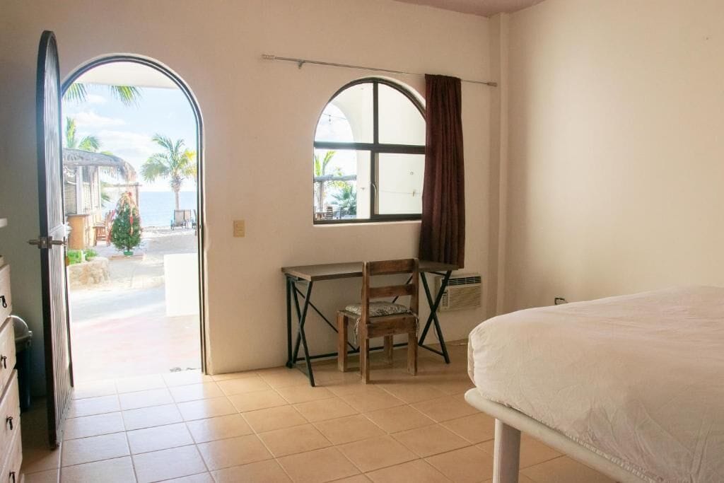 Una habitación con cama, escritorio y silla, ventana con cortinas y vista a la playa.