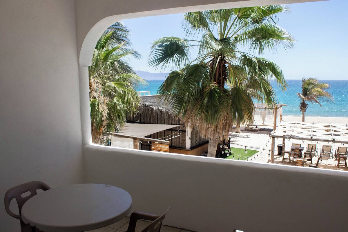Un balcon avec vue sur une plage, des palmiers et une enseigne indiquant « La Ventana ».