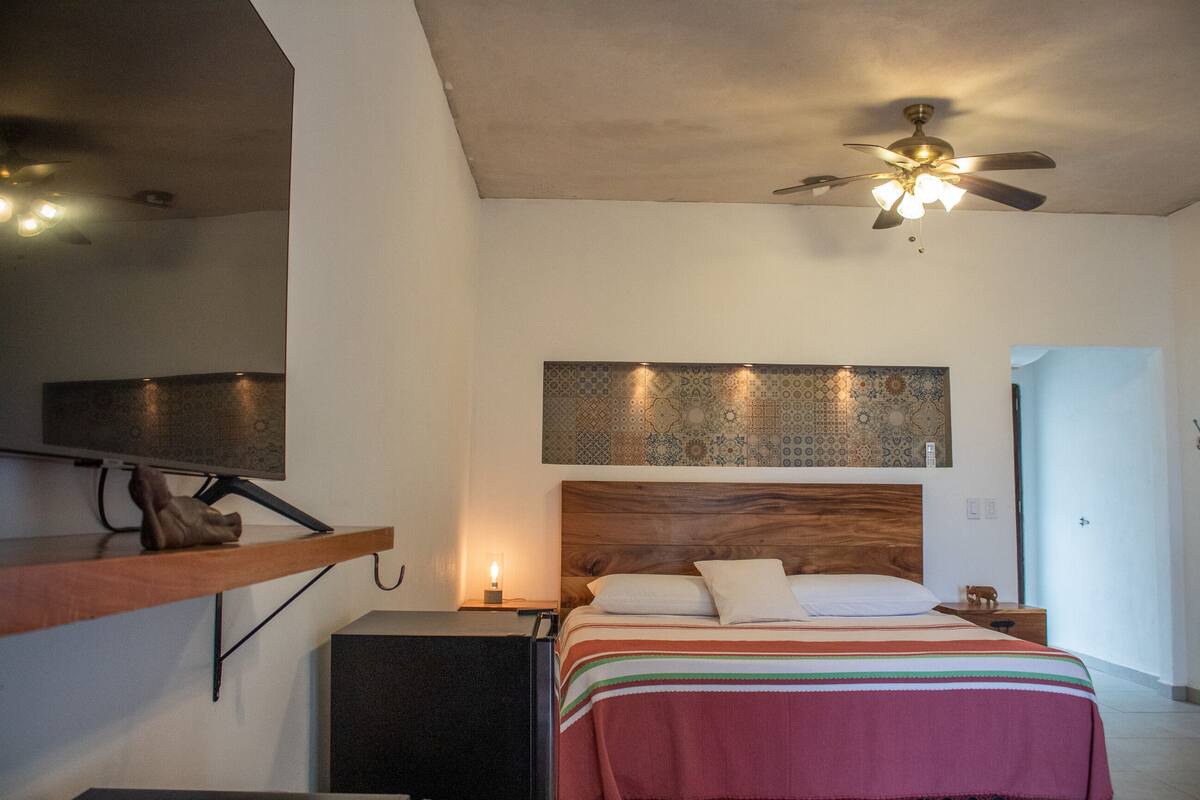 Une chambre à coucher comprenant un lit, une table de chevet, un téléviseur fixé au mur, un ventilateur de plafond et une décoration murale à motifs au-dessus du lit.