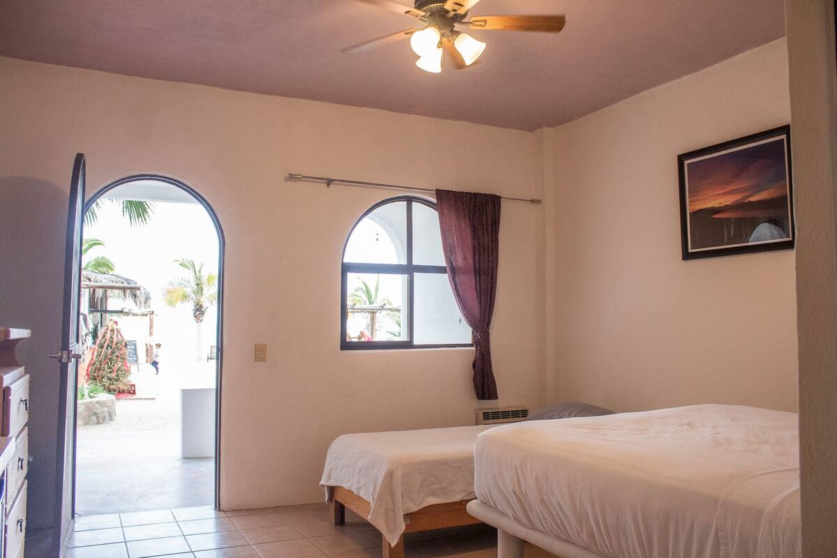 Une chambre avec deux lits, une fenêtre avec des rideaux, un ventilateur de plafond et une vue sur l’extérieur.