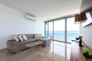 A modern living room with a grey sectional sofa, a small coffee table, and a large sliding glass door leading to a balcony with a view of the sea.