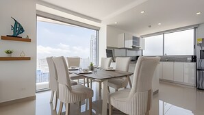 A modern dining area with a glass table and white chairs, a kitchen with white cabinets, and a view of a cityscape. - Awesome Sea View and Historic Center “Morros City” (Cartagena de Indias)