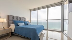 A modern bedroom with a large bed, a nightstand, a lamp, and a view of the ocean through large windows. - Awesome Sea View and Historic Center “Morros City” (Cartagena de Indias)
