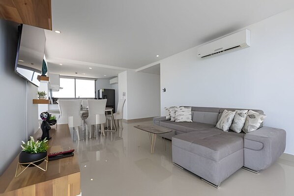 A modern living room with a grey sofa, a dining area with a table and chairs, and a flat-screen TV mounted on the wall.