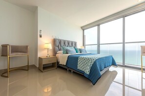 A modern bedroom with a large bed, bedside tables, a nightstand, and a view of the ocean through a large window.