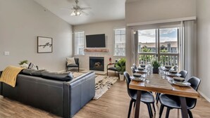 Ein modernes Wohnzimmer mit Kamin, einem Essbereich mit Holztisch und Stühlen sowie großen Fenstern mit Jalousien.