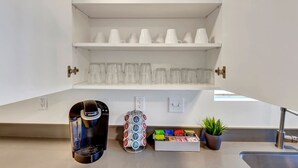 Een kitchenette met een koffiezetapparaat, een glazen kan, een houder voor koffiekapsules en een kleine potplant.