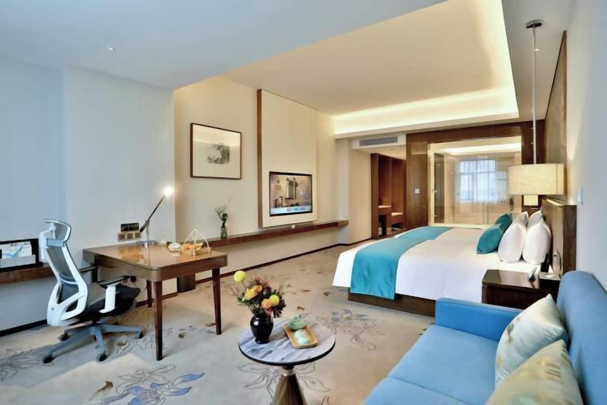 A hotel room with a bed, a desk, a chair, a sofa, a lamp, and a small table with flowers.