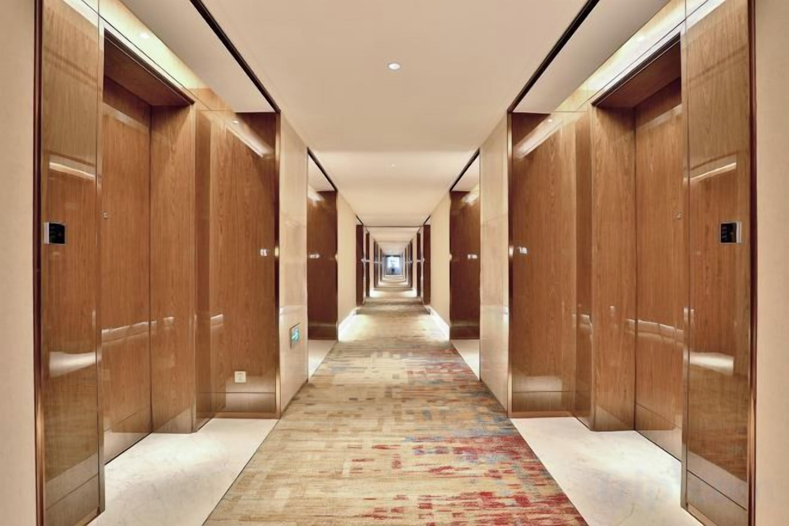 Un couloir d’hôtel avec des portes en bois, une moquette à motifs et un luminaire au plafond.